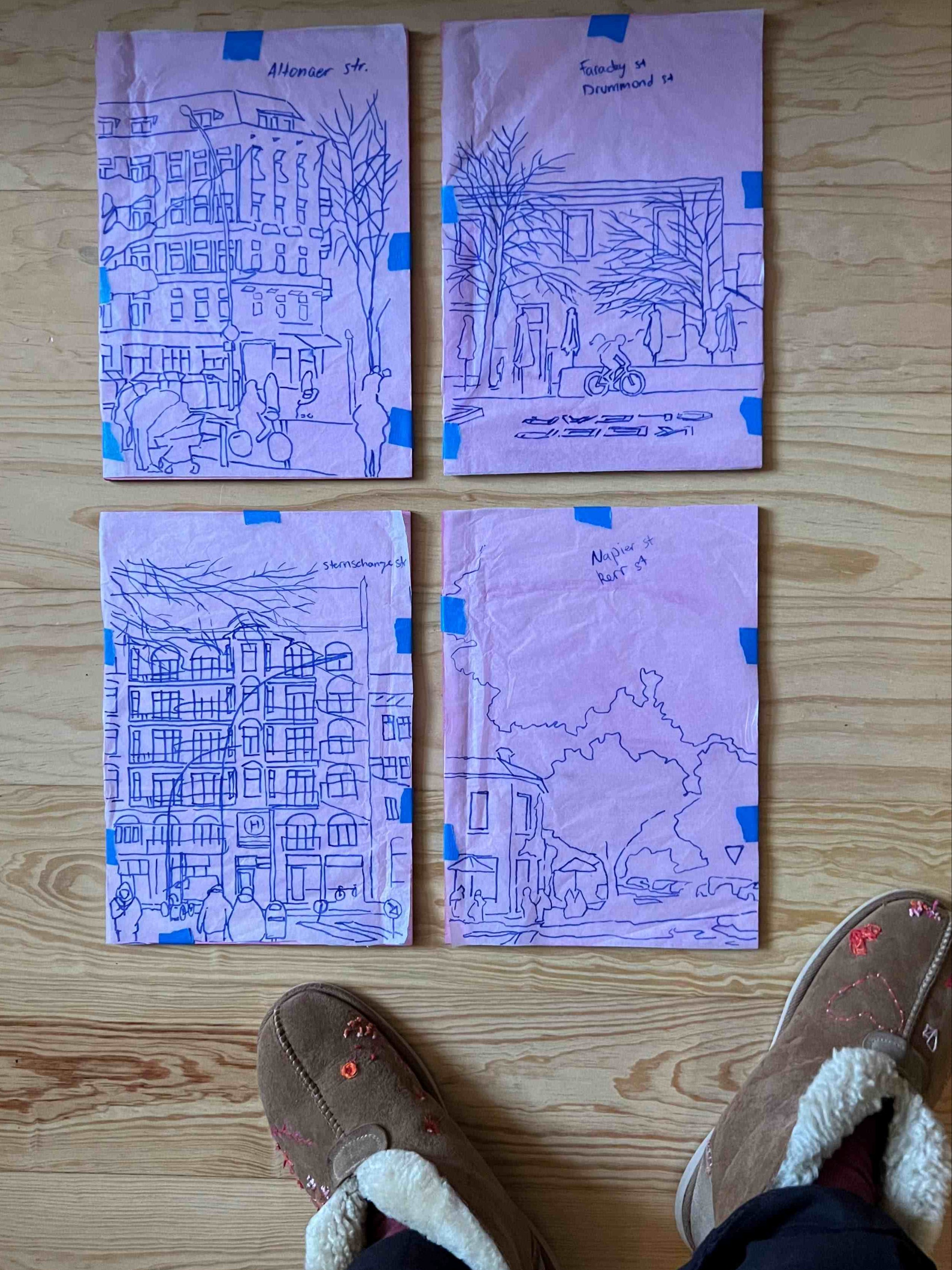 Four purple printed sheets with blue ink outlines of buildings on a wooden floor, with a person's feet visible.